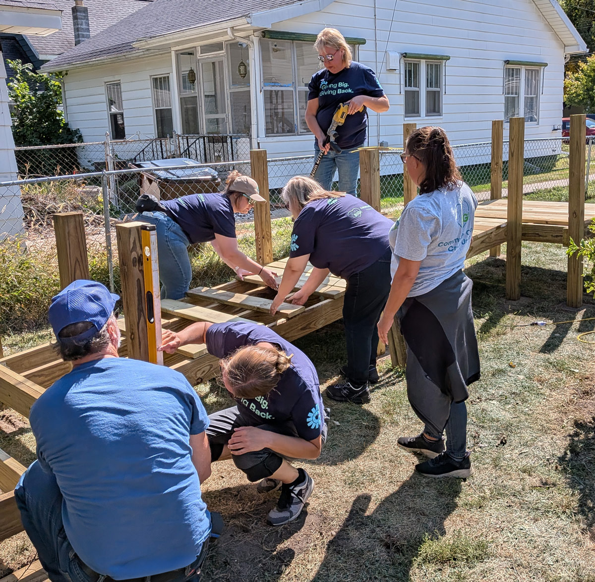 Volunteers assemble a wooden bed frame outdoors, working together with tools during a hands-on community service project.